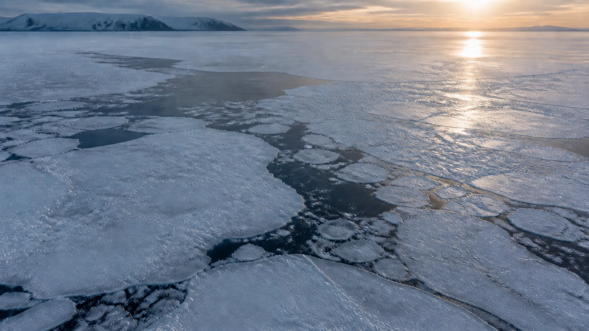 Oblique aerial view of coastal sea ice crossed by dark leads under low winter sunlight.