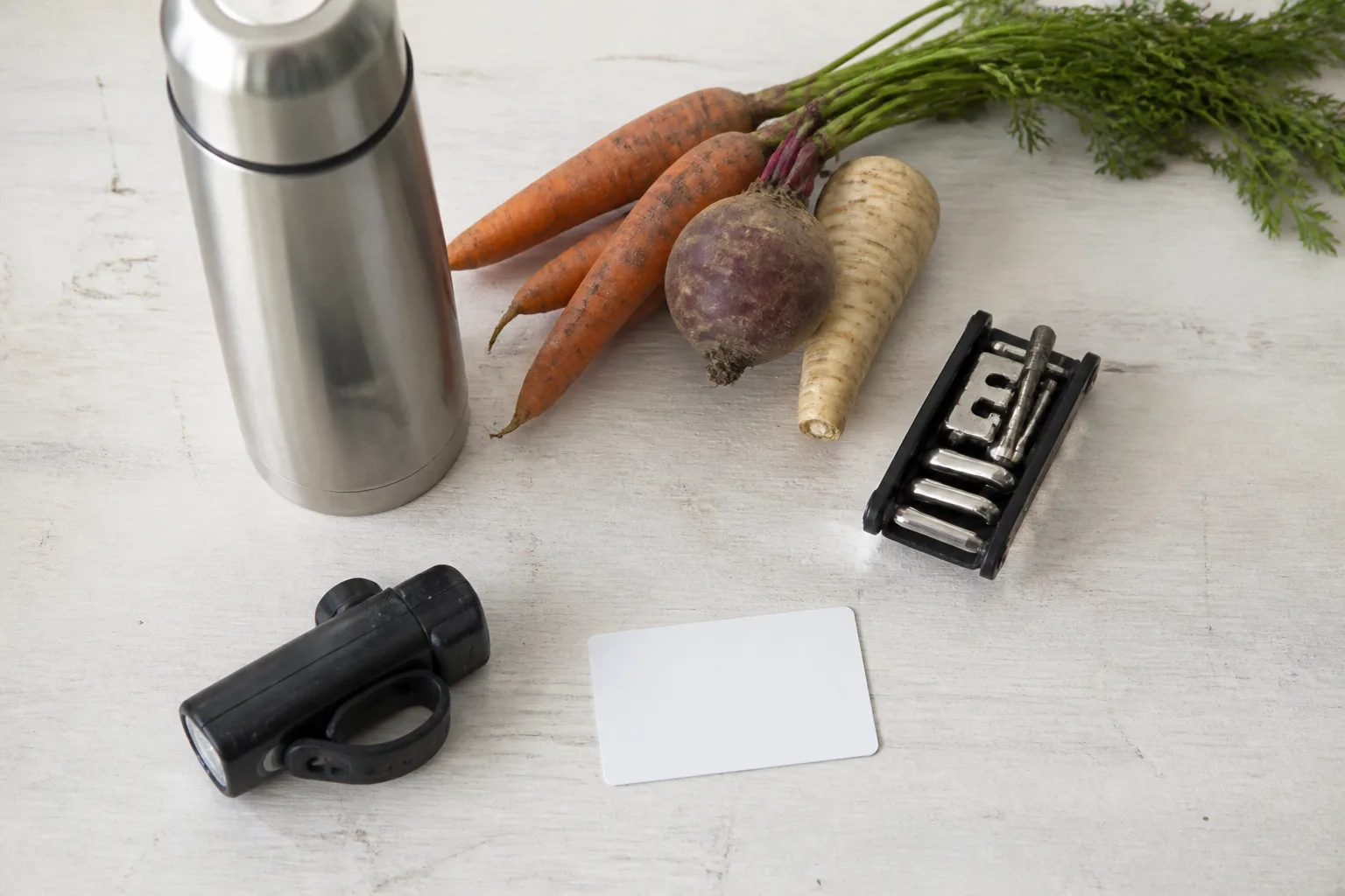 A bicycle light, transit card, vegetables, flask, and repair kit lie on a pale table.
